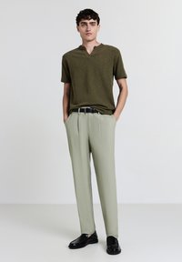 Giovane uomo in piedi con le mani in tasca, indossa una camicia a maniche corte verde oliva, pantaloni grigio chiaro, cintura nera e scarpe in pelle nera.