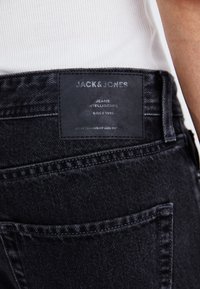 Jeans de mezclilla negra con una etiqueta de cuero con texto en relieve. Incluye un bolsillo trasero estándar y detalle de costura en contraste.