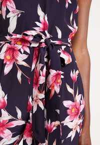 Combinaison fleurie bleu marine avec des fleurs roses et orange. Elle comporte une ceinture nouée à la taille et un tissu doux et léger avec un design fluide.