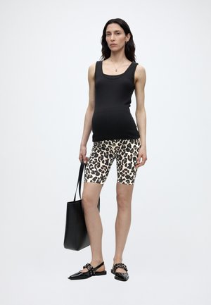 Femeie în picioare purtând un top negru fără mâneci, pantaloni scurți de biciclist cu imprimeu leopard, pantofi slingback negri cu ținte și ținând o geantă neagră mare.