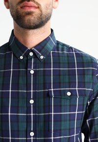 Camicia a scacchi in verde scuro, blu marino e bianco. Presenta un colletto a punta, bottoni bianchi e una tasca sul petto. Tessuto in cotone.
