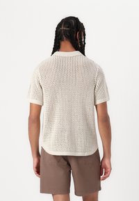 Rövid ujjú, nyitott hálós dizájnnal készült póló off-white színben, bordázott gallérral és mandzsettákkal, kerekített alsó szegéllyel, barna rövidnadrággal párosítva.