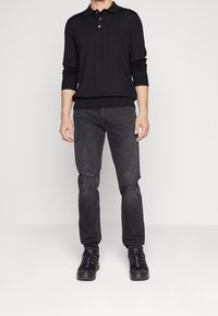 Man som bär en långärmad svart polotröja, slitna mörkgrå jeans och svarta sneakers, stående mot en enkel ljus bakgrund.