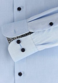 Helles blaues Hemd mit schwarzen Knöpfen, Knopfmanschetten-Details und gemustertem Stoff, der unter den gefalteten Manschetten hervorschaut. Glatte Textur.
