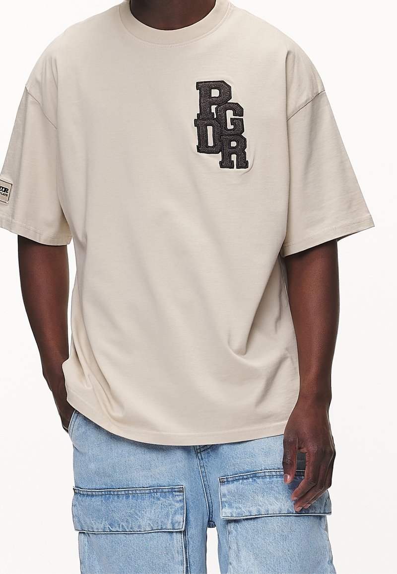 Camiseta de algodón beige con mangas cortas, que presenta un logotipo bordado en negro "PGDR" en el lado izquierdo del pecho. Textura suave, corte relajado.