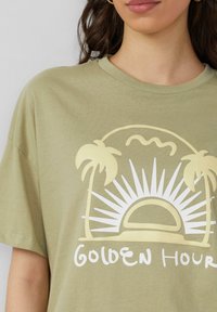Oliwkowa koszulka z krótkim rękawem, z żółtą grafiką palm i wschodzącego słońca, z napisem "ZŁOTA GODZINA" poniżej.