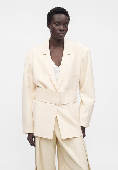 Γυναίκα με σκούρο δέρμα που φοράει υπερμεγέθη κρεμ σακάκι με φαρδιά ζώνη και αντίστοιχο παντελόνι με φαρδιά πόδια, στέκεται μπροστά σε λευκό φόντο.