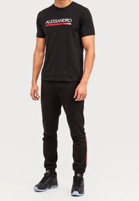 Camiseta de algodón negra con texto "ALESSANDRO" en blanco y rojo; combinada con joggers negros con rayas rojas y zapatillas deportivas negras.
