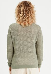 Maglione lavorato a maglia verde chiaro con motivo a rombi, polsini e orlo a coste. La texture appare aperta e traspirante.