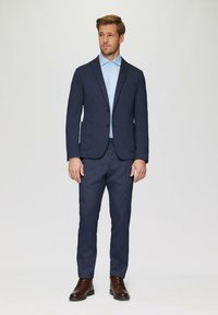 Marineblazer und passende Hose aus feinem Stoff. Hellblaues Hemd mit Kragen darunter. Braune Schnürschuhe runden das Outfit ab.