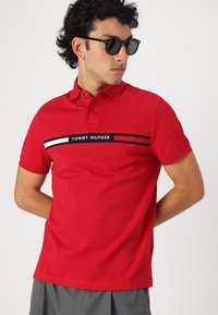 Rød polo t-shirt i bomuld med krave, korte ærmer og en vandret logo-stribe i hvid og sort tværs over brystet.