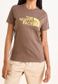 Hnědé bavlněné tričko s krátkým rukávem, které má na hrudi poškrábané žluté logo "THE NORTH FACE". Hladká textura.
