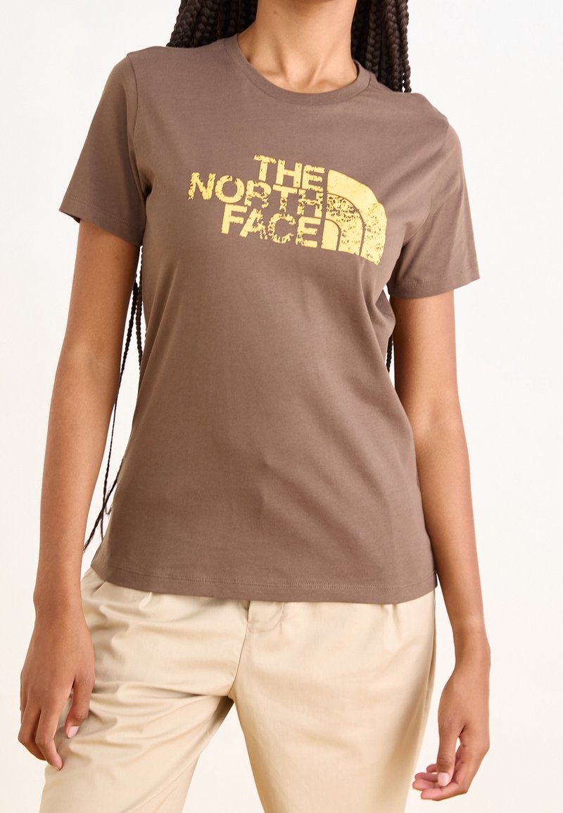 Hnědé bavlněné tričko s krátkým rukávem, které má na hrudi poškrábané žluté logo "THE NORTH FACE". Hladká textura.