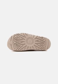 Εξωτερική σόλα παπουτσιού από μπεζ καουτσούκ με βαθιά ζιγκ-ζαγκ και κυκλικά σχέδια πέλματος και λογότυπο της μάρκας στο κέντρο, σε λευκό φόντο.