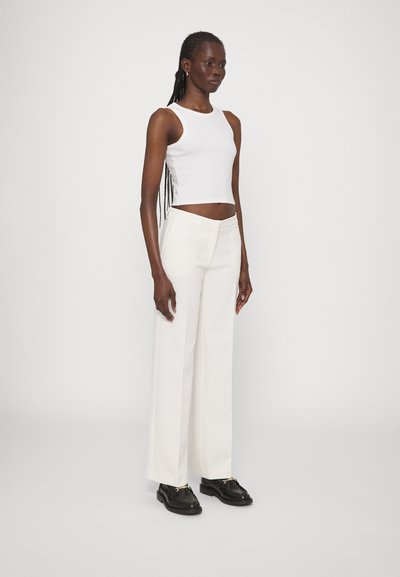 Un débardeur blanc court associé à un pantalon large crème taille haute. Des chaussures noires à semelles épaisses. Tissu lisse, design minimal.