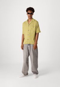 Chemise à manches courtes, boutonnée, de couleur jaune avec des rayures blanches, un pantalon en jean gris ample et des baskets blanches. Lunettes de soleil noires comme accessoire.