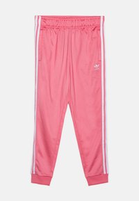 Różowe sportowe spodnie z białymi paskami po bokach, elastycznym pasem i ściągaczami na nogawkach. Materiał wydaje się gładki i lekki. Znakomita cecha to logo Adidas.