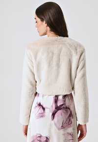 Veste en fausse fourrure beige raccourcie avec des manches longues, présentant une texture douce. Porteée sur une jupe blanche ornée de motifs floraux roses.