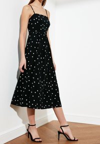 Kobieta w czarnej sukience midi w białe gwiazdki i cienkimi ramiączkami, w połączeniu z czarnymi sandałami na wysokim obcasie, stojąca na drewnianej podłodze.