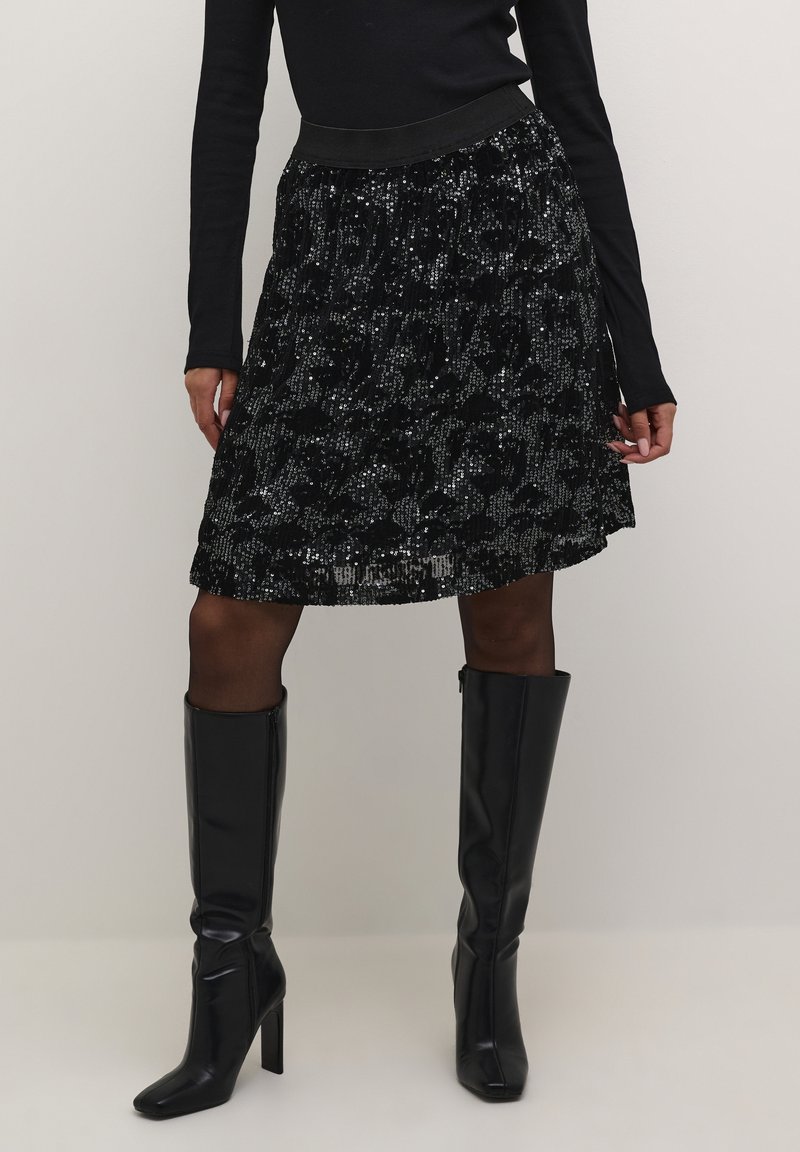 Femme portant une jupe noire à sequins longueur genou, un haut noir à manches longues, des collants noirs et des bottes noires à talons montant jusqu'au genou.