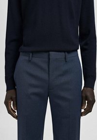 Mężczyzna noszący dopasowane ciemnoniebieskie eleganckie spodnie z szlufkami na pasek i czarną, wsuniętą w spodnie, koszulę z długim rękawem, z rękami spoczywającymi po bokach.