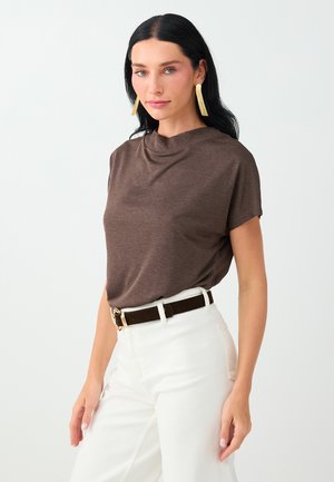 Femme aux longs cheveux noirs porte un haut marron à manches courtes, un pantalon blanc avec une ceinture noire, et de longues boucles d’oreilles dorées sur fond uni.