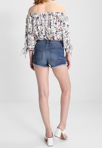 Valkoinen olkapäät paljastava kukkakuvioinen paita, jossa on pitkät hihat, yhdistetty sinisiin farkkushortseihin ja valkoisiin paksukorkoisiin sandaaleihin.