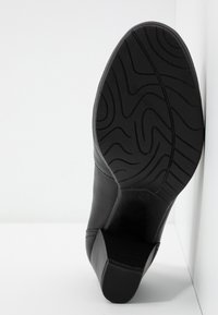 Semelle de chaussure à talon en cuir noir avec motif de bande de roulement texturé et ondulé, positionnée verticalement contre un mur d'angle blanc.