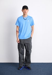 Modra Nike majica s logotipom, sivi cargo hlače z stranskimi žepi, črna kapa in črne superge z modrim podplatom. Oseba stoji na modrem preprogi.