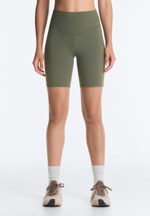 Femme portant un short cycliste taille haute vert olive, des chaussettes blanches montantes et des chaussures de sport beige, debout devant un fond blanc uni.