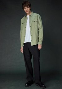 Light green button-up shirt with two chest pockets, worn over a white T-shirt. Black wide-leg pants and black sturdy shoes complete the outfit.