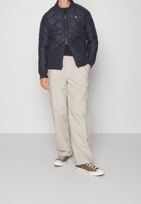 Granatowa pikowana kurtka z wysokim kołnierzem i zamkiem z przodu, w zestawieniu z beżowymi luźnymi spodniami i brązowymi trzewikami płóciennymi.