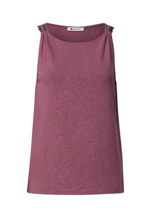 Femme blonde portant un haut mauve sans manches et un jean bleu, tenant un sac beige, debout légèrement tournée avec un fond neutre.