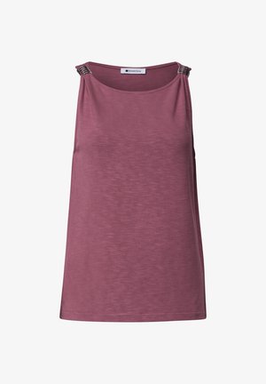 Femme blonde portant un haut mauve sans manches et un jean bleu, tenant un sac beige, debout légèrement tournée avec un fond neutre.
