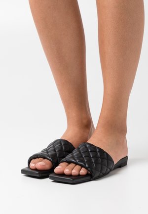 Sorte kviltede slidesandaler med flad sål, åben tå-design og en tekstureret overflade med diamantmønster. Glat finish og letvægtig.