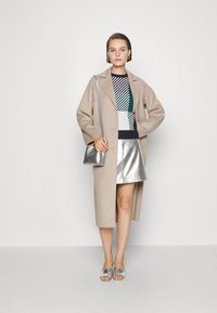 Beige lange jas over een multikleuren gebreide trui, metallic zilveren minirok en glanzende zilveren slides. Kleine zilveren crossbody tas.