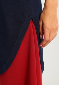Top lavorato a maglia blu navy con uno spacco laterale, che rivela uno strato rosso sottostante. Primo piano di una mano sul capo, che mette in mostra la trama e il contrasto di colore.