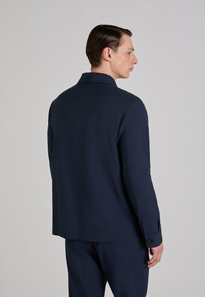 Mężczyzna w granatowej marynarce i spodniach stoi bokiem, głowa odwrócona, na tle jasnoszarego, jednolitego tła.