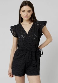 Combinaison en dentelle noire avec un décolleté en V, des manches courtes à volants et un lien à la taille. Le tissu présente des motifs découpés complexes.