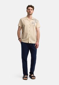 Beige kortærmet skjorte med dekorative blå mønstre, parret med marineblå bukser og sorte sandaler, hvilket skaber et afslappet outfit.