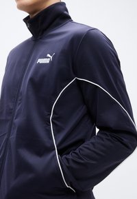 Μπλε ναυτικό τζάκετ με φερμουάρ, κατασκευασμένο από ομαλό ύφασμα, με ψηλό γιακά, λευκές λεπτομέρειες και λευκό λογότυπο Puma στην αριστερή πλευρά.