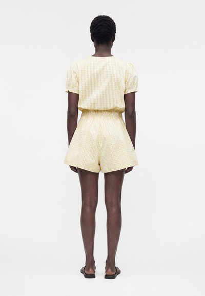 Γυναίκα που στέκεται με την πλάτη, φοράει ανοιχτό κίτρινο καρό τοπ με κοντά μανίκια και ταιριαστό σορτς με ψηλή μέση και μαύρα σανδάλια.