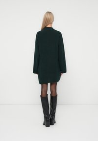 Zaļš adīts pārmērīgi liels džemperis, garas piedurknes, augsts dekoltē, vidusceļa garums, apvienots ar ceļgalussas melnām ādas zālēm. Gluda tekstūra.