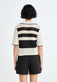 Hæklet top med sort og hvid horisontal striber, korte ærmer, bølgede kanter og en åben vævet tekstur. Matchet med sorte shorts.