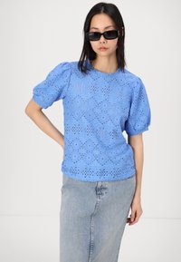 Top bleu clair à manches bouffantes en dentelle ajourée avec des découpes géométriques et un col rond, associé à une jupe en jean et des lunettes de soleil foncées.