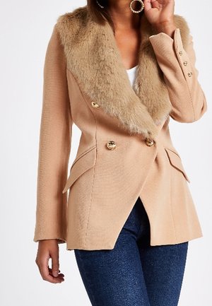 Mujer con un abrigo beige de doble botonadura con un amplio cuello de piel sintética y botones dorados, combinado con vaqueros de color azul oscuro.