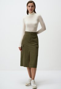 Olijfgroene katoenen rok met zijzakken, knie-lange split aan de voorkant, gecombineerd met een aansluitende witte coltrui en witte sneakers.