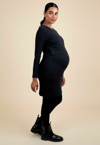 Nėščia moteris, vilkinti juodą ilgos rankovės suknelę, juodas leginsus ir juodus ankle batus, stovinti šonu prieš šviesiai smėlinį foną.