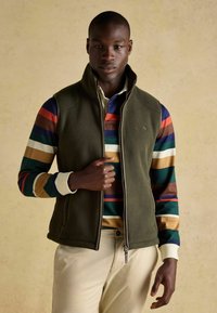 Zielona polarowa kamizelka z wysokim kołnierzem i zapięciem na zamek, noszona na kolorowej, w paski, długim rękawie. Model nosi beżowe spodnie.