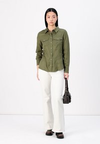 Camicia verde oliva con bottoni, dotata di due tasche sul petto, abbinata a pantaloni larghi bianchi e stivali marrone scuro. Il modello tiene in mano una piccola borsa nera.